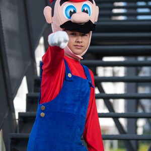 Op de afbeelding: Een kind verkleed als Super Mario, met een rode hoed met een witte 'M' en een rood shirt met lange mouwen en een blauwe overall. Het kind wijst met zijn rechterhand.