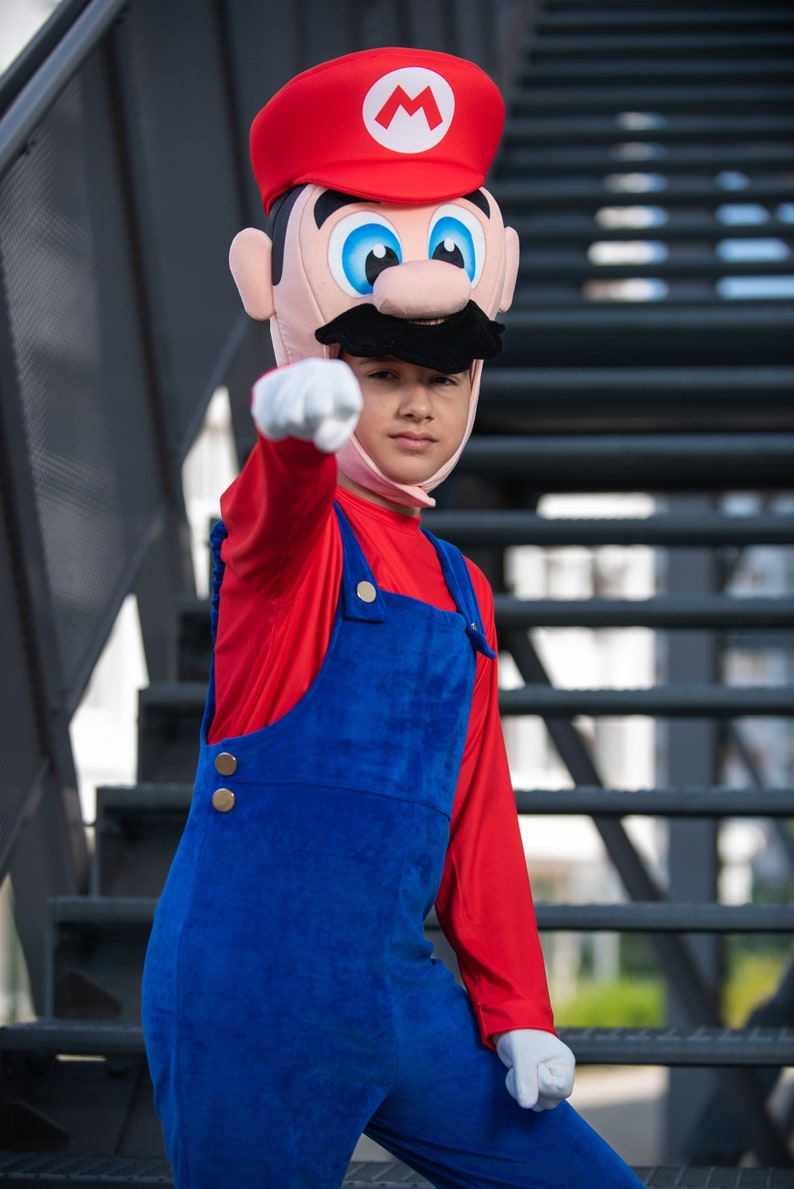 Op de afbeelding: Een kind verkleed als Super Mario, met een rode hoed met een witte 'M' en een rood shirt met lange mouwen en een blauwe overall. Het kind wijst met zijn rechterhand.