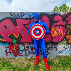 May include: A blue Captain America costume with a matching mask and shield. The shield is white, red, and blue with a white star in the centre. The costume includes red boots and is worn in front of a graffiti wall.