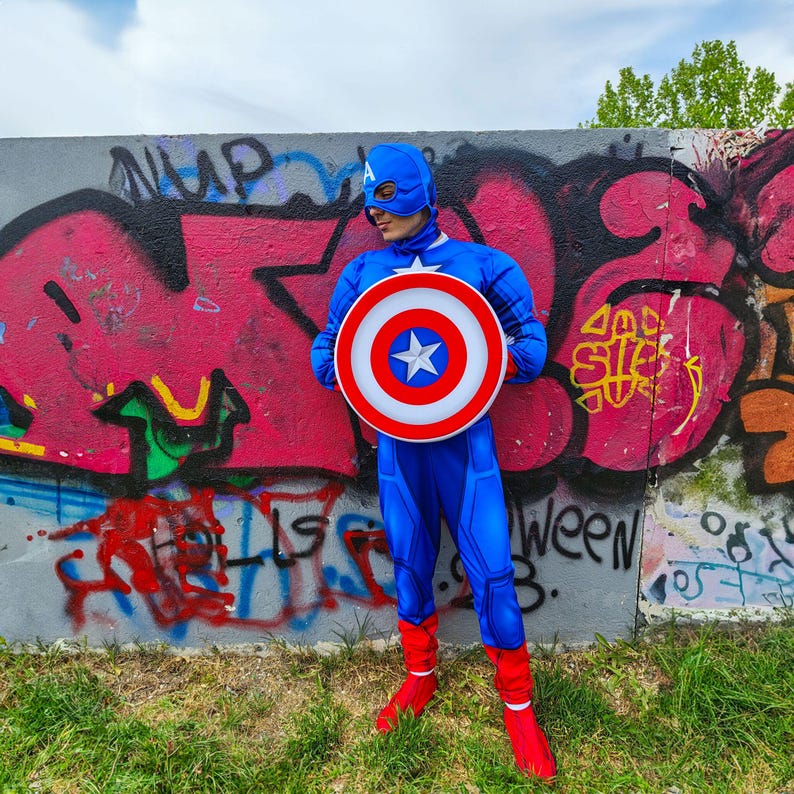 May include: A blue Captain America costume with a matching mask and shield. The shield is white, red, and blue with a white star in the centre. The costume includes red boots and is worn in front of a graffiti wall.