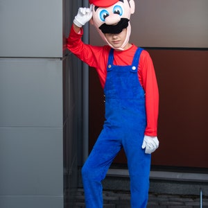 Op de afbeelding: Een kind verkleed als Super Mario, met een rood shirt, een blauwe overall en een rood-witte muts met een snor. Het kind staat naast een grijze muur.