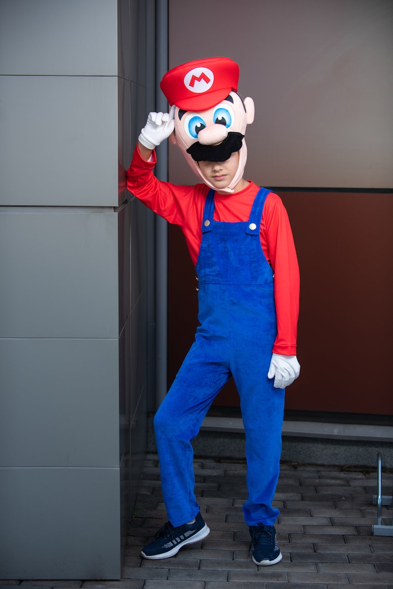 Op de afbeelding: Een kind verkleed als Super Mario, met een rood shirt, een blauwe overall en een rood-witte muts met een snor. Het kind staat naast een grijze muur.