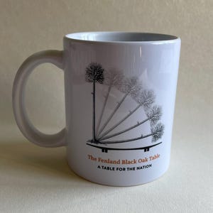 May include: White ceramic mug with a curved handle. The mug features a black and white illustration of a tree with multiple branches and the text "The Fenland Black Oak Table" and "A Table for the Nation" in orange.
