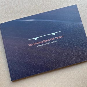 May include: A book titled "The Fenland Black Oak Project: A Table for the Nation" on a textured dark blue background. The cover features a minimalist design with a white line drawing and text in white and red.