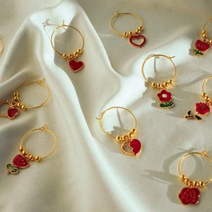 Peut inclure: Neuf boucles d'oreilles créoles dorées avec des breloques en forme de cœur rouge, de fleur et de rose. Les breloques sont attachées aux créoles par de petites perles dorées.