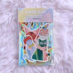 May include: A pack of "Cute Cat" stickers featuring whimsical illustrations of cats in various disguises. The stickers are packaged in a holographic sleeve with text that reads "Cats in Disguise Stickers Pack" and "Studio Cty". The cats are depicted in colorful outfits.