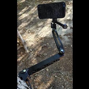 May include: A black articulated phone mount with a clamp base and a pivoting arm, featuring the text LONGSHOT ARCHERY printed in gold on the side. The device holds a smartphone in a horizontal position, secured by a tightening knob at the top.