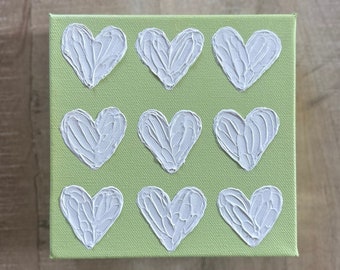 May include: A green canvas painting with nine white textured hearts arranged in a grid pattern.