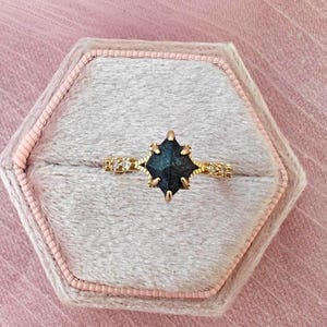 May include: A gold ring with a dark blue gemstone in a starburst setting. The ring is displayed in a pink velvet jewellery box. The band features small decorative accents. The background is a soft pink fabric.