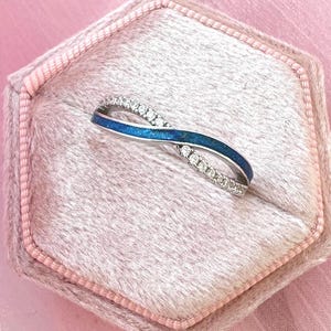 May include: An elegant silver ring with a blue enamel inlay and a row of small, clear gemstones. The ring is in an infinity shape and is displayed in a pink velvet ring box against a pink background.