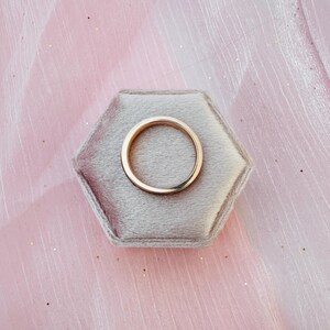 May include: A gold wedding band rests on a gray velvet hexagonal ring box. The ring is a simple, polished band with a smooth surface. The background is a soft pink fabric.