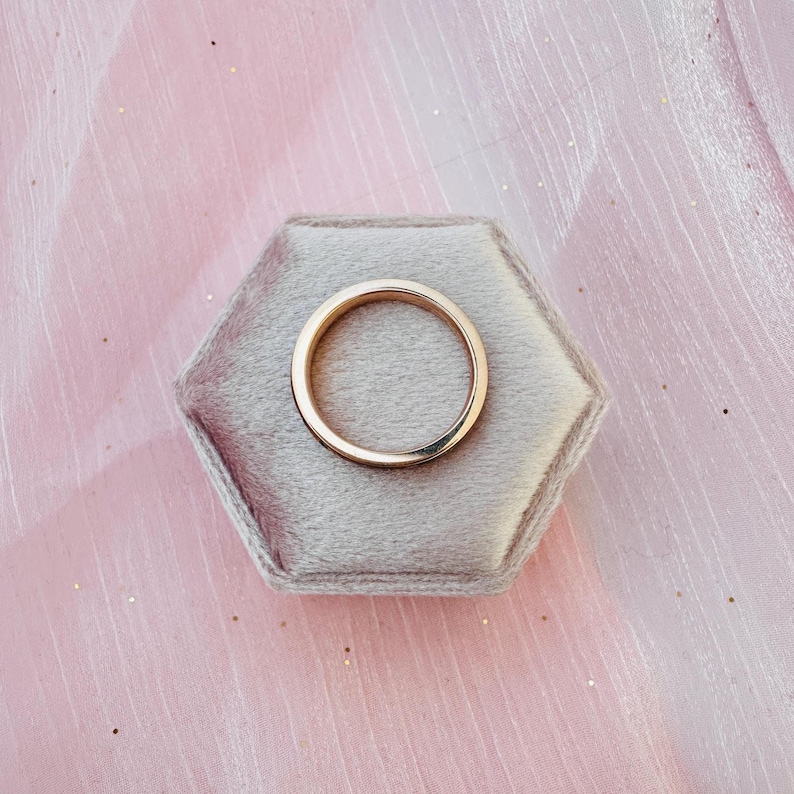 May include: A gold wedding band rests on a gray velvet hexagonal ring box. The ring is a simple, polished band with a smooth surface. The background is a soft pink fabric.