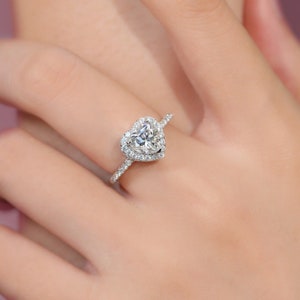 May include: A silver ring featuring a heart-shaped diamond, encircled by a halo of smaller diamonds. The ring's band is also set with small diamonds. The ring is displayed on a finger. The ring's design is elegant and eye-catching.