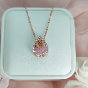 May include: A delicate rose gold necklace with a teardrop-shaped pendant. The pendant features a pink and gold resin design with a clear crystal halo.