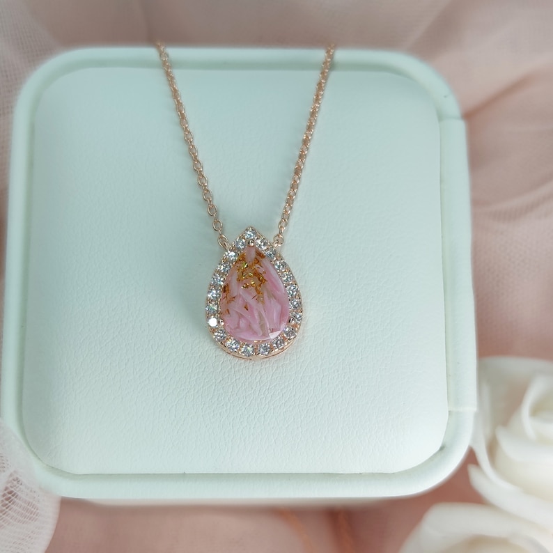 May include: A delicate rose gold necklace with a teardrop-shaped pendant. The pendant features a pink and gold resin design with a clear crystal halo.