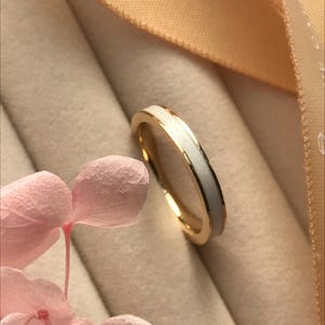 May include: A gold ring with a white enamel inlay. The ring is set against a soft, neutral background with a pink flower in the foreground. The ring's design is simple and elegant.