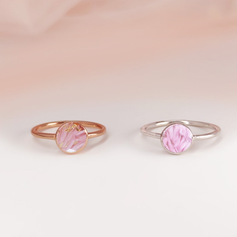 May include: Two delicate rings with pink floral designs encased in clear resin. The ring on the left has a rose gold band, while the ring on the right has a silver band.