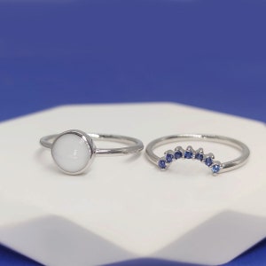 May include: Two silver rings on a white surface. One ring has a white stone and the other has a row of blue stones.