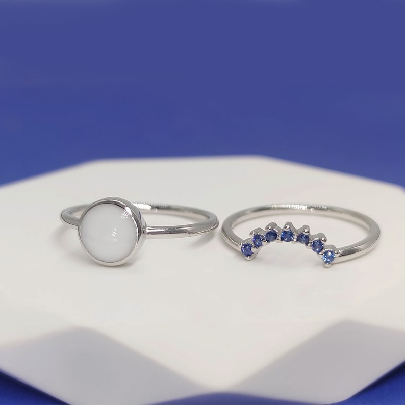 May include: Two silver rings on a white surface. One ring has a white stone and the other has a row of blue stones.