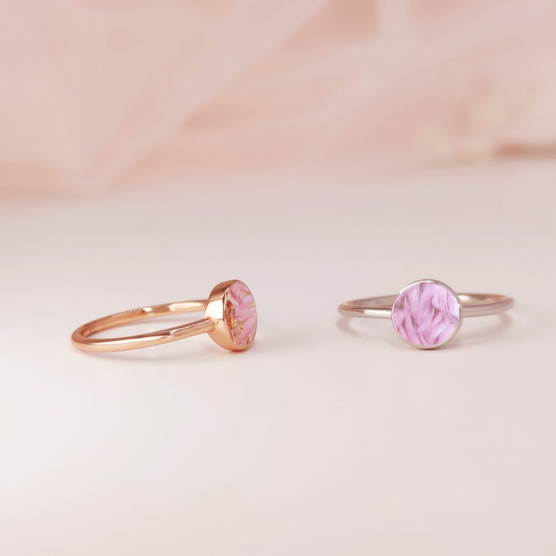May include: Two delicate rings with pink floral resin inlays. The ring on the left is gold, and the ring on the right is silver.