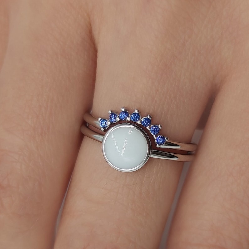 May include: A silver ring with a white oval stone and a row of blue sapphires around the stone.