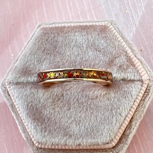 May include: A gold-toned ring with a band filled with red, gold, and green flecks. The ring is set on a gray velvet ring box. The ring's design is visible and detailed.