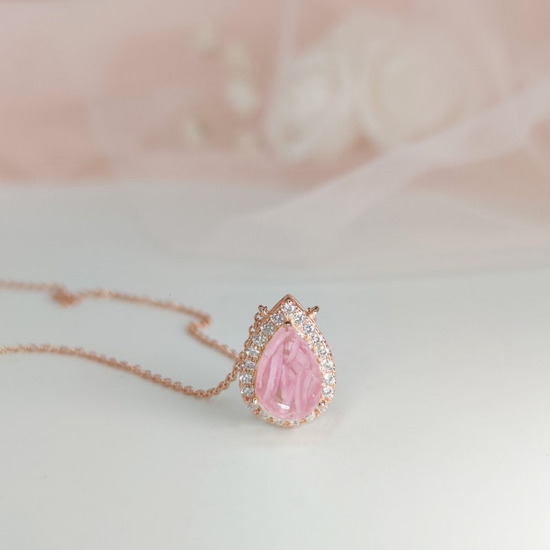 May include: A delicate rose gold necklace with a pear-shaped pink gemstone pendant surrounded by a halo of sparkling clear stones.