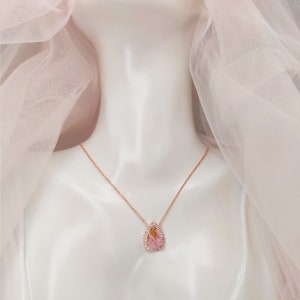 May include: A delicate rose gold necklace with a teardrop-shaped pendant featuring a pink gemstone surrounded by smaller clear stones.