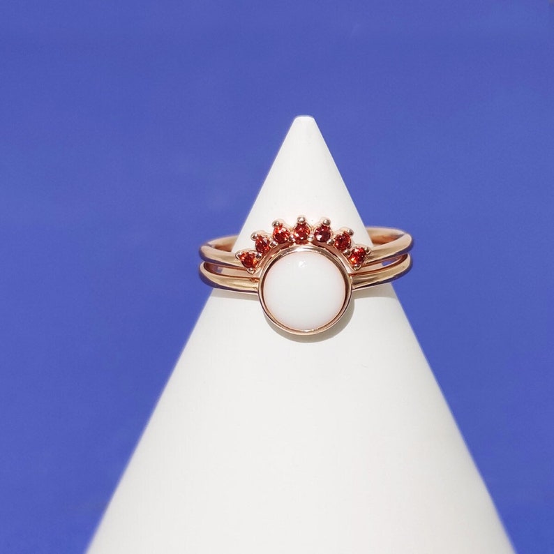 May include: A pair of rose gold rings with a white stone and red gemstones. The rings are stacked on top of each other and are on a white display stand.
