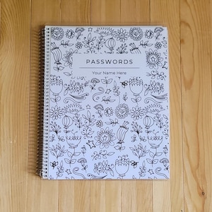 May include: A spiral-bound notebook with a white cover featuring a black and white floral design. The cover has the word "PASSWORDS" and "Your Name Here" printed on it. The notebook is on a wooden surface.