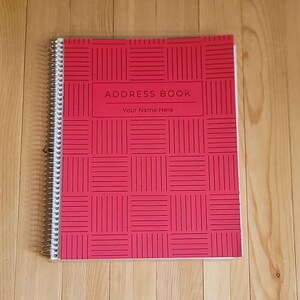 May include: Red address book with a grid pattern and spiral binding. The cover features the text "ADDRESS BOOK" and "Your Name Here". The notebook is on a wooden surface.