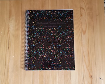 May include: A spiral-bound journal with a black cover featuring a colorful pattern of small squares in red, blue, green, and yellow. The text on the cover reads My Diamond Painting Journal and Your Name Here in a simple, dark font.
