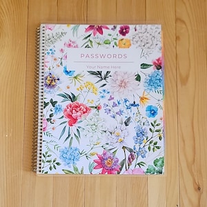 May include: A spiral-bound notebook with a floral design featuring pink, blue, and yellow flowers. The cover reads "PASSWORDS" with space for a name. The notebook is on a wooden surface.