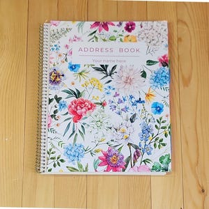 May include: A spiral-bound address book with a floral design. The cover features a white background with watercolor illustrations of various flowers in shades of pink, blue, yellow, and purple. The text "ADDRESS BOOK" and "Your name here" are visible.
