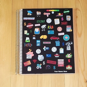 May include: Black spiral-bound notebook with colorful financial-themed stickers. Stickers include dollar signs, a piggy bank, a car, and the words "Due Today," "Money," and "Paid." The bottom of the notebook has the text "Your Name Here."