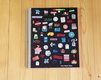 May include: A spiral-bound notebook with a black cover featuring a variety of colorful financial-themed stickers, including icons of coins, a piggy bank, a car, and text labels like DUE TODAY, MONEY, PAID, DEBT FREE, and Your Name Here.