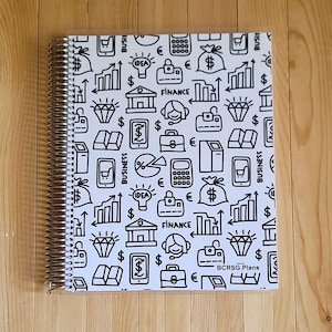 May include: A spiral-bound planner with a white cover featuring a repeating black doodle pattern of financial icons, including money bags, charts, and diamonds. The text "BCRSG Plans" is visible in the lower right corner. The planner is on a light brown wooden surface.