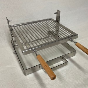 May include: A stainless steel portable grill with a square cooking surface. It features a grid of parallel bars and two wooden handles. The grill is designed for outdoor cooking and grilling.