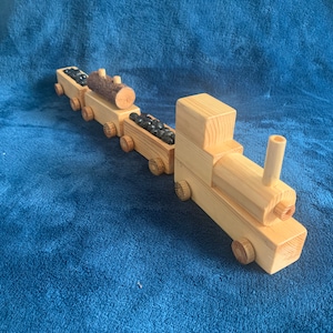 May include: A wooden toy train with three cars. The engine car has a chimney and a log of wood in the back. The middle car is filled with black beads and the last car has a log of wood in the front. The train is on a blue surface.