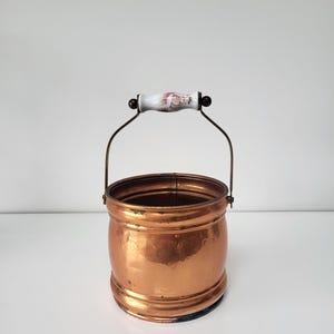 May include: A copper bucket with a decorative handle. The bucket has a rounded shape with a wide base and a handle made of a white ceramic with a floral design. The handle is attached to the bucket with a metal wire.