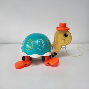 May include: A vintage pull-along toy turtle. It has a teal shell with yellow and white shapes, orange feet, and a yellow head with a small orange hat. A yellow string extends from the turtle's head. The toy is designed for young children.