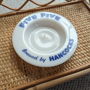 May include: White ceramic ashtray with blue lettering that reads "Five Five Brewed by Hancocks".