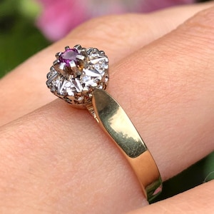 May include: A gold ring with a central purple gemstone encircled by smaller, clear stones. The ring has a plain gold band. The background is blurred, showing green leaves and a pink flower. The ring is a classic design.