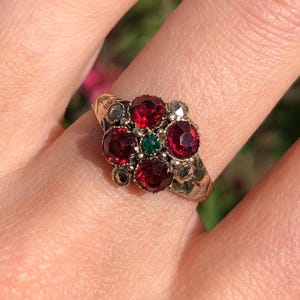 May include: A gold ring featuring a cluster of red gemstones arranged in a floral pattern, with a small green gemstone at the center. The ring has a detailed, textured band.