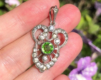 May include: A silver-toned pendant with a heart-shaped design, encrusted with clear and green gemstones. The central green stone is round, surrounded by smaller clear stones. The pendant has a bail for attaching to a chain.