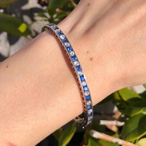 May include: A silver bracelet with alternating blue and white gemstones. The bracelet is worn on a wrist, showcasing the design and sparkle of the gemstones. The gemstones are set closely together, creating a continuous line of color.