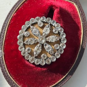 May include: A round, antique brooch with a central floral design. The brooch features numerous clear, sparkling stones set in a silver or platinum metal. It rests in a red velvet-lined case with a brown border, suggesting a vintage jewelry piece.