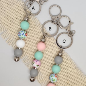 May include: Three keyrings with silver hardware and beaded accents. Each keyring features a combination of pastel-coloured beads, including mint green, pink, white, and grey, with floral patterned beads. The keyrings are displayed on a neutral fabric.