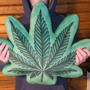 Peut inclure: Un coussin en peluche vert en forme de feuille de marijuana. Le coussin a un contour vert foncé et des nervures vert clair.