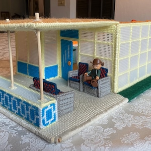 Op de afbeelding: Een miniatuur huis van plastic canvas met een blauwe en witte buitenkant, een bruin dak en een wit balkon. Het huis heeft een blauwe deur en een klein figuurtje van een persoon die op een stoel op het balkon zit.